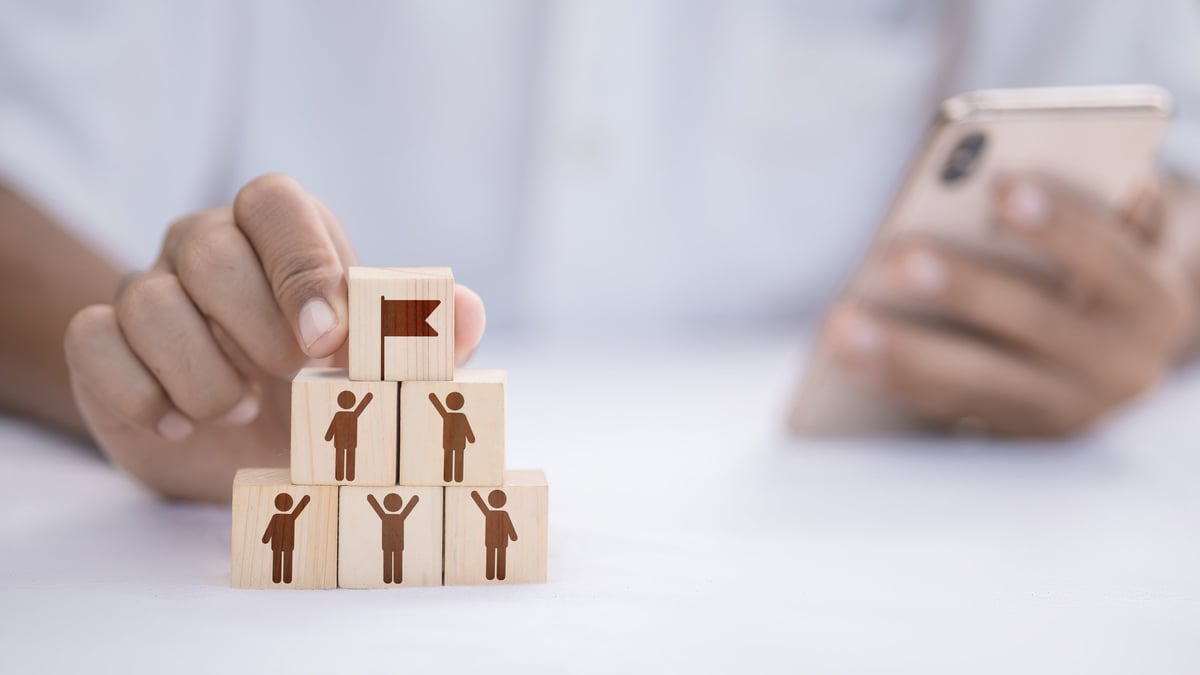 Hand stacking wooden blocks into pyramid with people icons and red flag on top, concept of leadership, team building, goal achievement, human resources management, and employee motivation.